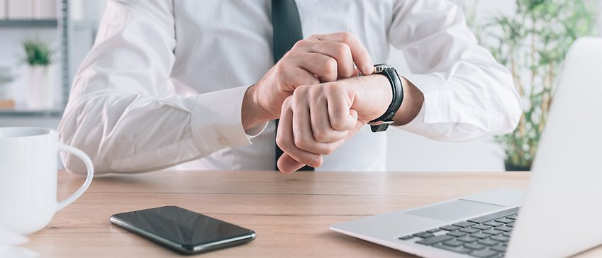 En närbild på en mans överkropp i skjorta och slips. Mannen tittar på sin klocka. På bordet finns en kopp, mobil och en dator.